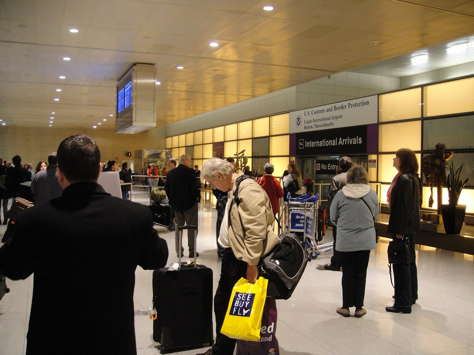 Travelers waiting in the International Arrivals hall at Boston Logan airport beneath a U.S. Customs and Border Protection sign